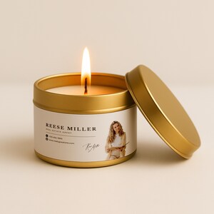 May include: A lit gold candle with a lid resting beside it. The candle has a white label with the name "Reese Miller, Real Estate Agent" and a photo of a woman. The candle is in a gold tin.