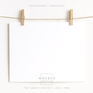 May include: A blank white rectangular paper is suspended by two wooden clothespins from a thin, light brown rope. The paper is centered, with the text "PHOTOSHOP • PHOTOPAE" at the top and "MOCKUP HAUS CO" and "PSD SMART OBJECT • JPG • PNG" at the bottom.