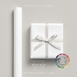Puede incluir: Una caja de regalo blanca con una cinta y lazo plateados, junto a un rollo de papel blanco. La imagen incluye el texto "Photoshop Photopea" y "Mockup Haus Co."