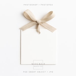 May include: A blank, off-white square tag with a beige ribbon tied in a bow at the top. The tag has a small hole near the top for hanging. The words "PHOTOSHOP • PHOTOPEA" are at the top, and "MOCKUP HAUS CO" and "PSD SMART OBJECT • JPG" are at the bottom.
