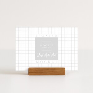 May include: A white card with a grid pattern and the text "Just Add Art" is displayed in a wooden holder. The card also features the text "MOCKUP MADE CO" and "JPG &bull; PSD". The overall aesthetic is minimalist and clean.