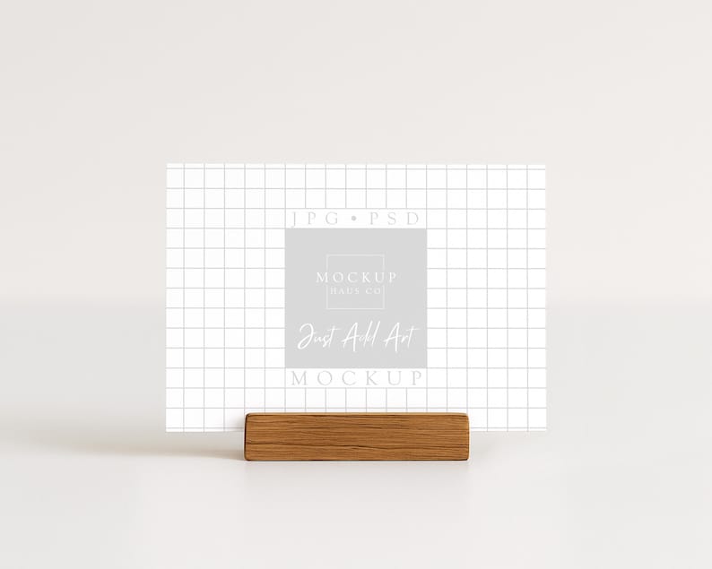 May include: A white card with a grid pattern and the text "Just Add Art" is displayed in a wooden holder. The card also features the text "MOCKUP MADE CO" and "JPG &bull; PSD". The overall aesthetic is minimalist and clean.