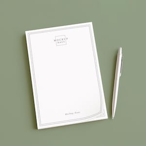 May include: A white notepad with a black border and the text "MOCKUP HAUS" is next to a silver pen on a green surface. The notepad has a simple, elegant design, and the pen is sleek and modern.
