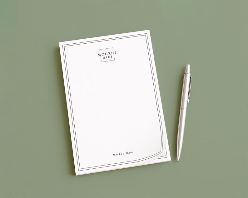 May include: A white notepad with a black border and the text "MOCKUP HAUS" is next to a silver pen on a green surface. The notepad has a simple, elegant design, and the pen is sleek and modern.