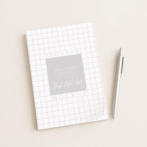May include: A white notebook with a grid pattern and the text "Just Add Art" is next to a silver pen. The notebook has a gray square with the text "Mockup Days Co" and "Mockup" on it.