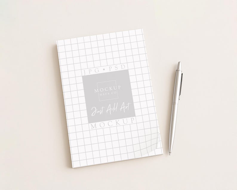 May include: A white notebook with a grid pattern and the text "Just Add Art" is next to a silver pen. The notebook has a gray square with the text "Mockup Days Co" and "Mockup" on it.