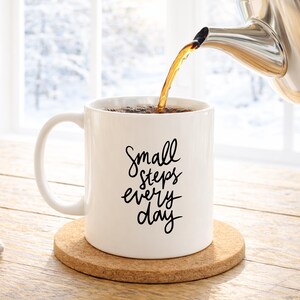 May include: A white ceramic mug with the text "Small steps every day" in black script is being filled with tea from a stainless steel teapot. The mug sits on a round cork coaster, with a snowy window in the background.