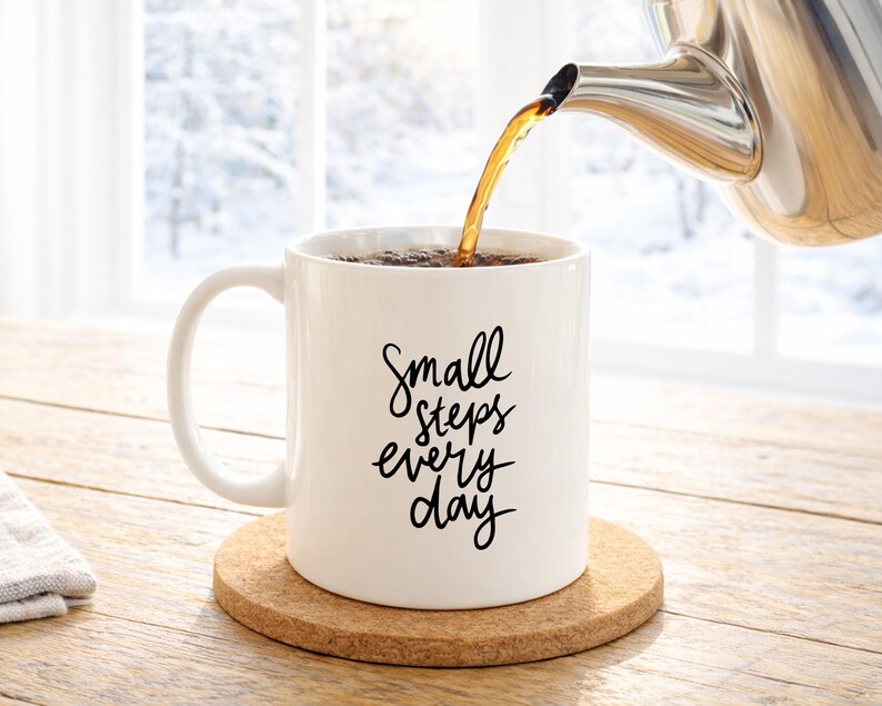 May include: A white ceramic mug with the text "Small steps every day" in black script is being filled with tea from a stainless steel teapot. The mug sits on a round cork coaster, with a snowy window in the background.