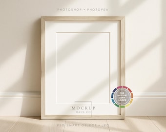 May include: A rectangular wooden picture frame with a white mount, leaning against a wall. The frame has a light wood grain finish. The text "MOCKUP HAUS CO" is visible at the bottom. The text "PHOTOSHOP • PHOTOPEA" is at the top.