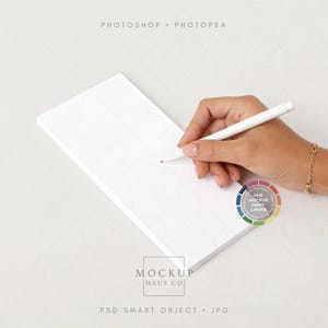 May include: A white notepad with a grid pattern is held by a hand, with a white pen poised above it. The image includes the text "Photoshop Photpea" and "Mockup Haus Co". A gold bracelet is on the wrist.