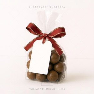 Puede incluir: Una bolsa transparente llena de caramelos de chocolate redondos y marrones, atada con una cinta roja y dorada y una etiqueta blanca en blanco. La bolsa está sellada en la parte superior.