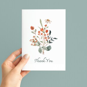 May include: A white card with a watercolour floral design, featuring orange and green flowers and foliage. The words "Thank You" are written in a delicate script at the bottom of the card.