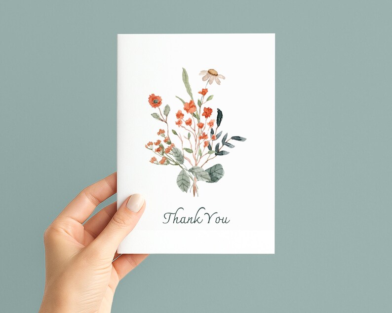 May include: A white card with a watercolour floral design, featuring orange and green flowers and foliage. The words "Thank You" are written in a delicate script at the bottom of the card.