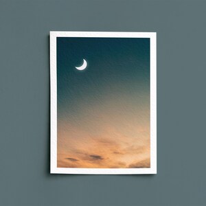 May include: A framed print featuring a crescent moon in a gradient sky. The upper portion is a deep teal, transitioning to orange and yellow hues at the bottom, suggesting a sunset or sunrise. The print has a white border.