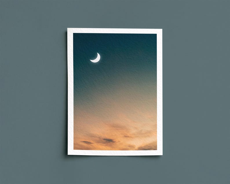 May include: A framed print featuring a crescent moon in a gradient sky. The upper portion is a deep teal, transitioning to orange and yellow hues at the bottom, suggesting a sunset or sunrise. The print has a white border.