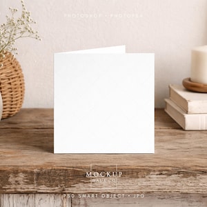 Op de afbeelding: Een blanco, witte wenskaart staat open op een rustiek houten oppervlak. De kaart heeft een subtiel ruitpatroon. Een kaars, boeken en een vaas met bloemen staan op de achtergrond, wat een zachte, neutrale esthetiek creëert.