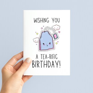 May include: A white birthday card with the text "WISHING YOU A TEA-RIFIC BIRTHDAY!" features a cartoon tea bag with a smiling face, clouds, and hearts. The card is held by a hand.