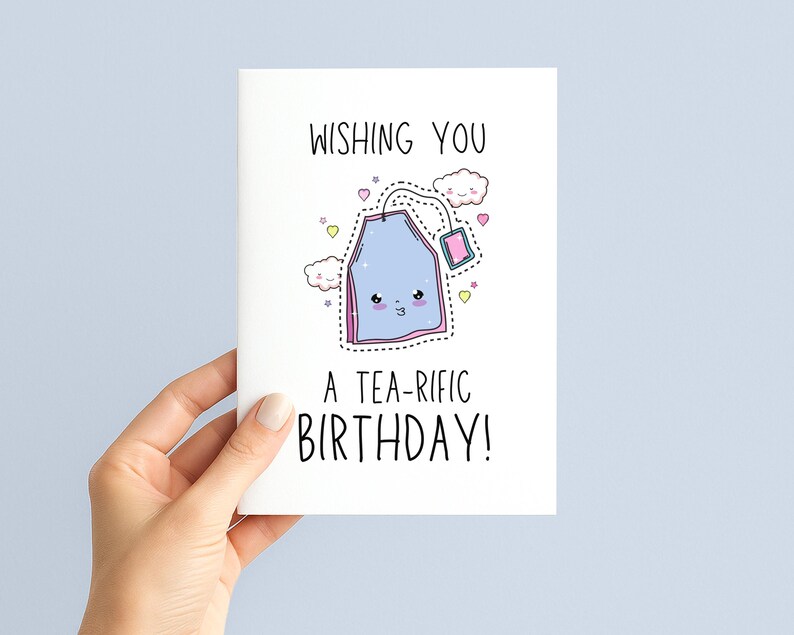 May include: A white birthday card with the text "WISHING YOU A TEA-RIFIC BIRTHDAY!" features a cartoon tea bag with a smiling face, clouds, and hearts. The card is held by a hand.