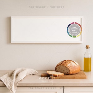 May include: A white rectangular picture frame hangs above a countertop with a loaf of bread, a cutting board, a bottle of oil, and a linen napkin. The bread is partially sliced, and the oil bottle has a cork stopper.