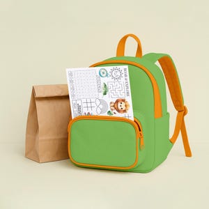 May include: A green backpack with orange accents and straps, a front pocket, and a zipper. A paper with a lion illustration and the text "ROAR & EXPLORE" is in the front pocket. A brown paper lunch bag is next to the backpack.