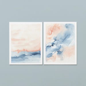 May include: Two framed watercolor paintings. The left painting features a soft peach and blue landscape. The right painting has abstract blue and peach watercolor washes. Both paintings are in white frames, set against a light blue background.