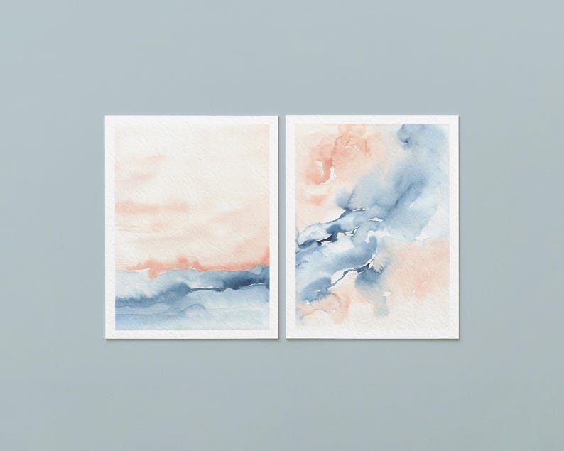 May include: Two framed watercolor paintings. The left painting features a soft peach and blue landscape. The right painting has abstract blue and peach watercolor washes. Both paintings are in white frames, set against a light blue background.