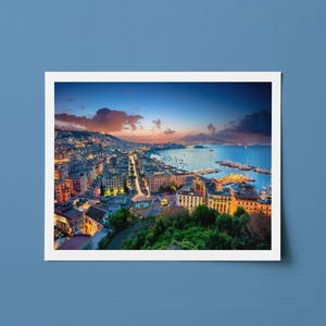 Puede incluir: Fotograf&iacute;a de una ciudad costera al anochecer, con edificios iluminados por farolas y un cielo colorido. La imagen est&aacute; enmarcada en un borde blanco, sobre un fondo azul.