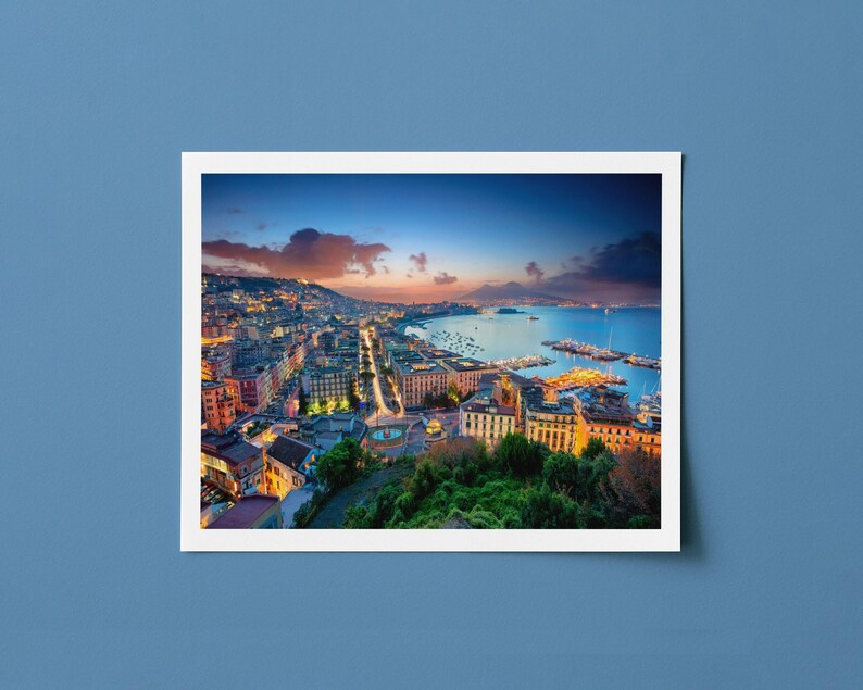Puede incluir: Fotograf&iacute;a de una ciudad costera al anochecer, con edificios iluminados por farolas y un cielo colorido. La imagen est&aacute; enmarcada en un borde blanco, sobre un fondo azul.