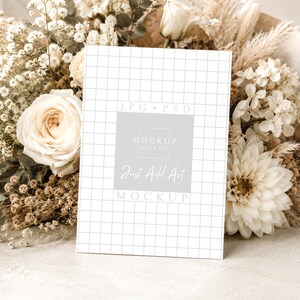 Pu&ograve; includere: Una carta bianca con un motivo a griglia e la scritta "Just Add Art Mockup" &egrave; appoggiata a un bouquet di fiori secchi e rose bianche. La carta ha un quadrato grigio chiaro con la scritta "Mockup Haus Co."