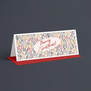 Pu&ograve; includere: Un biglietto di Natale piegato con base rossa e fronte bianco. Il fronte presenta un motivo di decorazioni natalizie dorate, rosse e nere. Le parole "Merry Christmas!" sono scritte in rosso.