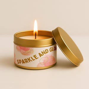 May include: A lit gold candle in a round tin with the lid off. The candle has a white label with pink floral designs and the words "Sparkle and Glow" in gold lettering.
