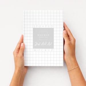 K&ouml;nnte beinhalten: Ein wei&szlig;es Buch mit einem Rastermuster wird von zwei H&auml;nden gehalten. Das Buch hat ein graues Quadrat mit dem Text "Mockup Haus Co" und "Just Add Art". Der Hintergrund ist wei&szlig;.