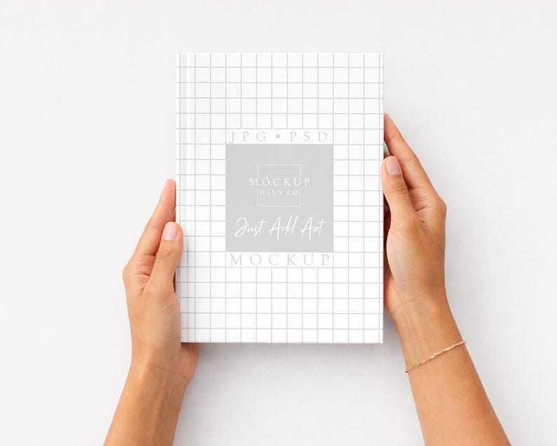 K&ouml;nnte beinhalten: Ein wei&szlig;es Buch mit einem Rastermuster wird von zwei H&auml;nden gehalten. Das Buch hat ein graues Quadrat mit dem Text "Mockup Haus Co" und "Just Add Art". Der Hintergrund ist wei&szlig;.