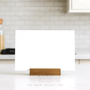 May include: A blank white card in a wooden holder, ideal for displaying messages or artwork. The holder is made of light brown wood. The background features a kitchen setting with white tile and a glass bottle.