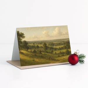 May include: A greeting card featuring a landscape painting of rolling green hills, trees, and a cloudy sky. A red Christmas ornament and a sprig of evergreen are next to the card.