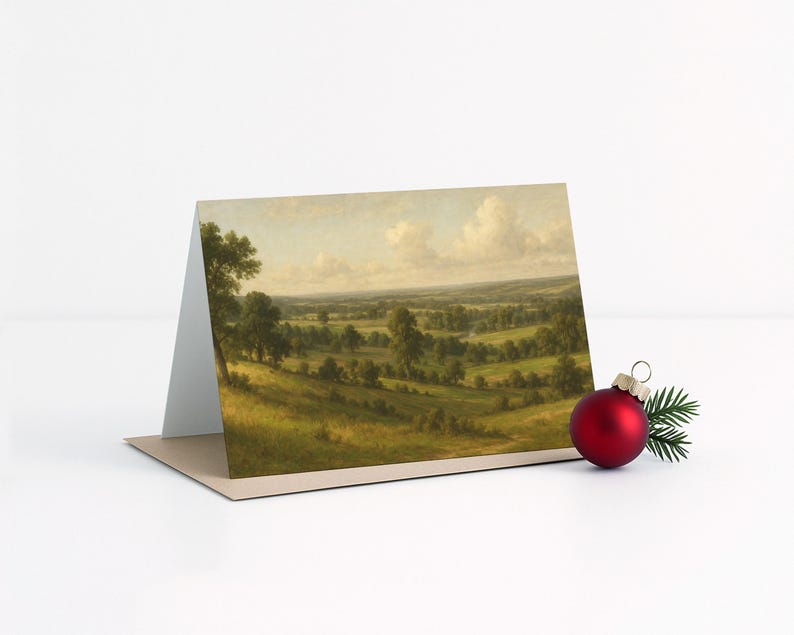 May include: A greeting card featuring a landscape painting of rolling green hills, trees, and a cloudy sky. A red Christmas ornament and a sprig of evergreen are next to the card.