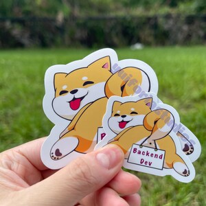 Op de afbeelding: Twee gestanste stickers met een cartoon Shiba Inu-hond in een ontspannen pose. De grotere sticker is een volledig lichaamsbeeld, terwijl de kleinere een bord heeft met de tekst "Backend Dev". De stickers zijn geel, wit en zwart.