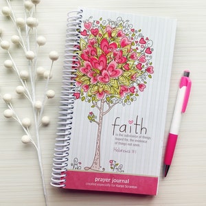 May include: A pink and white prayer journal with a spiral binding. The cover features a colorful illustration of a tree with hearts and the text "faith is the substance of things hoped for, the evidence of things not seen. Hebrews 11:1". The journal is titled "prayer journal" and says "created especially for Karen Scranton".