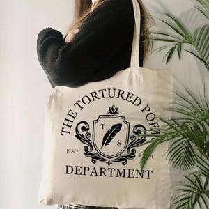 Puede incluir: Bolso tote de color crema con texto negro y un gráfico. El texto dice "THE TORTURED POETS DEPARTMENT" en un diseño circular, con "EST 2024" debajo. Un gráfico de escudo con una pluma está en el centro. El bolso tiene correas largas.