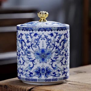 May include: A cylindrical ceramic jar with a white base and blue floral patterns. The lid features a gold-coloured lotus-shaped knob. The body displays intricate scrolling vines and a central flower motif containing the Chinese character for good fortune.