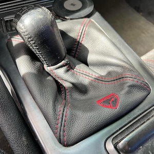 May include: Black leather shift boot with red stitching and a red emblem. The shift knob is black and textured. The boot is installed in a car's center console.