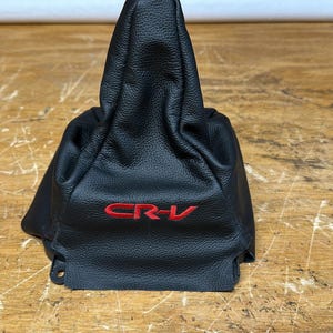 May include: Black leather shift boot with red "CR-V" lettering. The boot is designed to cover the gear shift lever in a vehicle. The leather has a textured appearance and is shaped to fit around the lever.