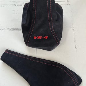 May include: Black suede car shift boot and console cover with red stitching. The shift boot has "VR-4" embroidered in red. The items are likely for a vehicle's interior.