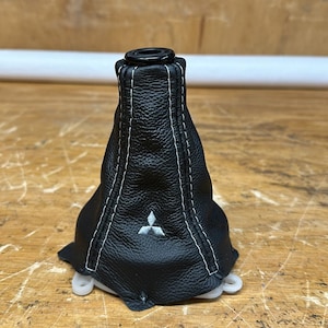 May include: Black leather shift boot with white stitching and a small emblem. The boot is designed to fit around a gear shift lever. The boot is sitting on a wooden surface.