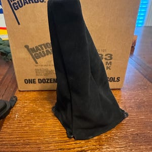 May include: A black, suede-like fabric cover, possibly for a car's gear shift, is the primary focus. The cover is draped over a wooden surface, with a cardboard box labeled "NATIONAL GUARDS" in the background.