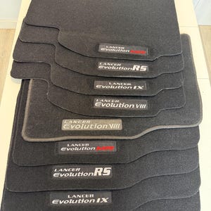 May include: A stack of black car floor mats. Each mat has a rectangular shape with a custom fit. The mats feature the "Lancer Evolution" logo in white and red, with "RS", "IX", and "VIII" model designations.