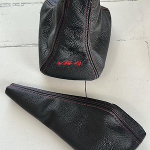 May include: Two black leather car shift boots with red stitching. The larger boot has the text "VR-4" in red. The leather has a textured appearance. One boot is upright, and the other is lying flat.