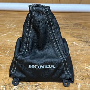 May include: Black leather Honda shift boot with white stitching. The boot has a textured surface and the Honda logo is visible. The shift boot is designed to fit a vehicle's gear shift.