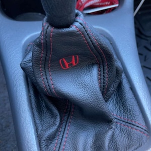 May include: Black leather shift boot with red stitching and a red Honda logo. The boot is designed to fit around a car's gear shift lever. The stitching provides a contrasting detail to the black leather.