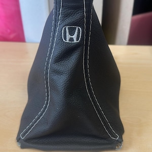 May include: Black leather shift boot with white stitching and the Honda logo. The boot has a textured surface and is designed to fit a vehicle's gear shift lever. The stitching provides a contrasting detail.